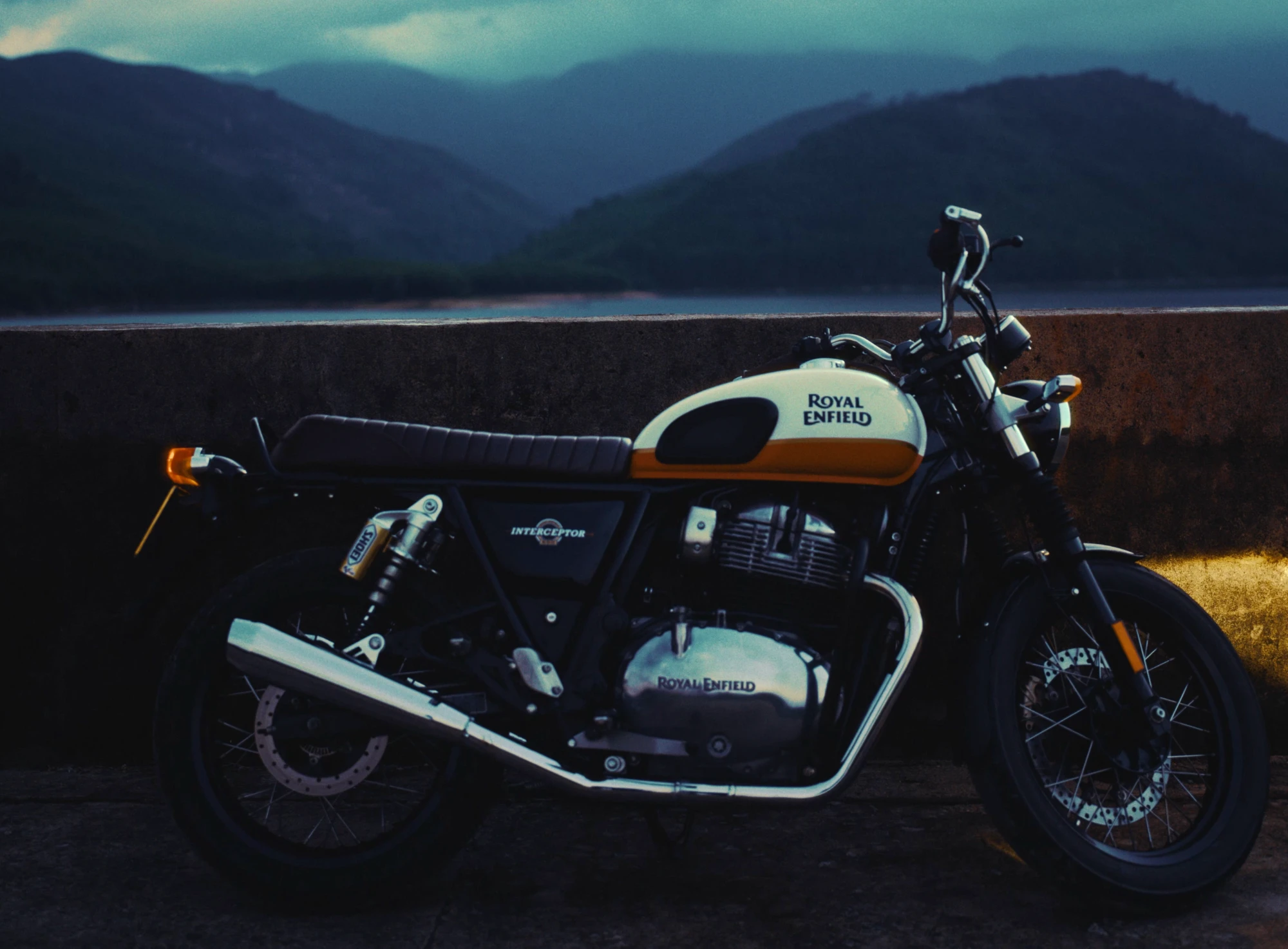 A rider and a Royal Enfield by a mountain lake at dusk, a helmet on the wall, the river from the air